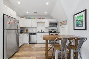 Une cuisine moderne avec des meubles blancs, des appareils en acier inoxydable, une table à manger en bois et un tableau encadré au mur.