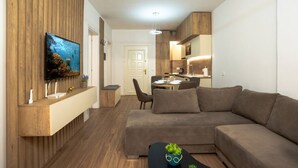Una sala moderna con un sofá seccional grande, un televisor de pantalla plana montado en un panel de madera, un comedor con mesa y sillas, y una kitchenette con encimera y armarios.