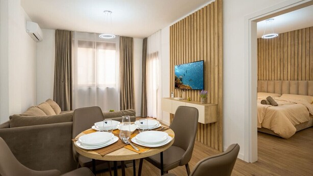 Una habitación de hotel moderna con comedor, televisor montado en una pared de paneles de madera, y un dormitorio separado con cama y cabecera.