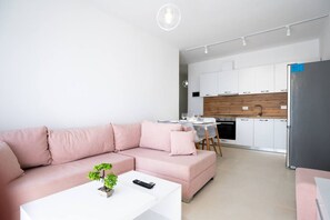 A modern living room with a pink sofa, a small white table with a plant and remote control, and a kitchen in the background with white cabinets and a wooden countertop.
