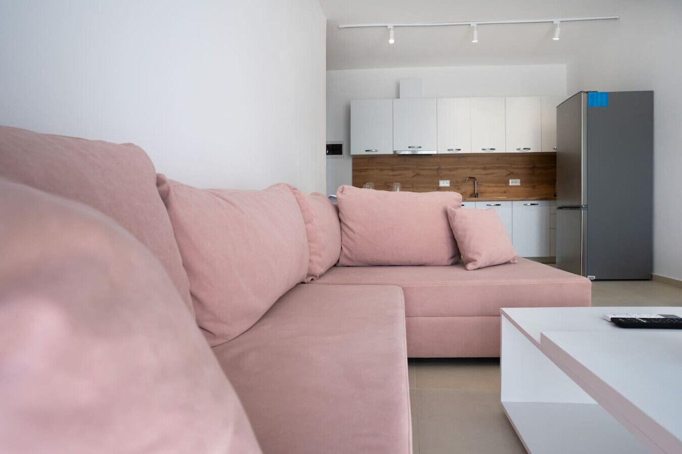Ein modernes Wohnzimmer mit einem rosafarbenen Sofa, einem weißen Couchtisch und einer Küchenzeile mit weißen Schränken und einem grauen Kühlschrank.