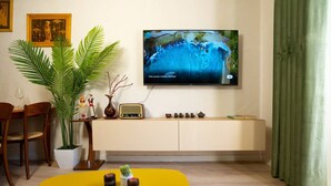 Ein Wohnzimmer mit Fernseher an der Wand, einem Esstisch, Stühlen, einer Topfpflanze, einer Vase, einem Radio und einer Kommode mit Tassen.