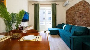 Ein gemütliches Wohnzimmer mit einem Holzespeisetisch, gedeckt für zwei Personen, einem blaugrünen Sofa, einer Ziegelwand und einem großen Fenster mit grünen Vorhängen.