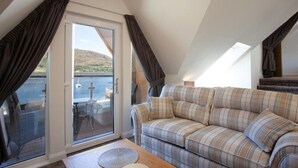 A cozy room with a sofa, a table, and a sliding glass door leading to a balcony with a view of a body of water and hills. - Loch View Apartment in Fort William Town Centre (Fort William)