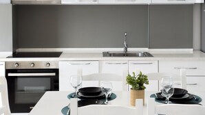A modern kitchen with white cabinets, a built-in oven, a sink, and a dining table set for two with black plates and glasses. - Terrace View Apartment Durres (Durrës)
