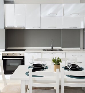A modern kitchen with white cabinets, a built-in oven, a sink, and a dining table set for two with black plates and glasses. - Terrace View Apartment Durres (Durrës)