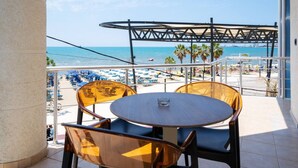 Un balcon avec une table et des chaises donnant sur une plage parsemée de parasols et de palmiers.
