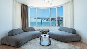 Un salon moderne avec deux canapés gris, un pouf à motifs, une petite table ronde et une vue sur un pont enjambant une étendue d’eau.