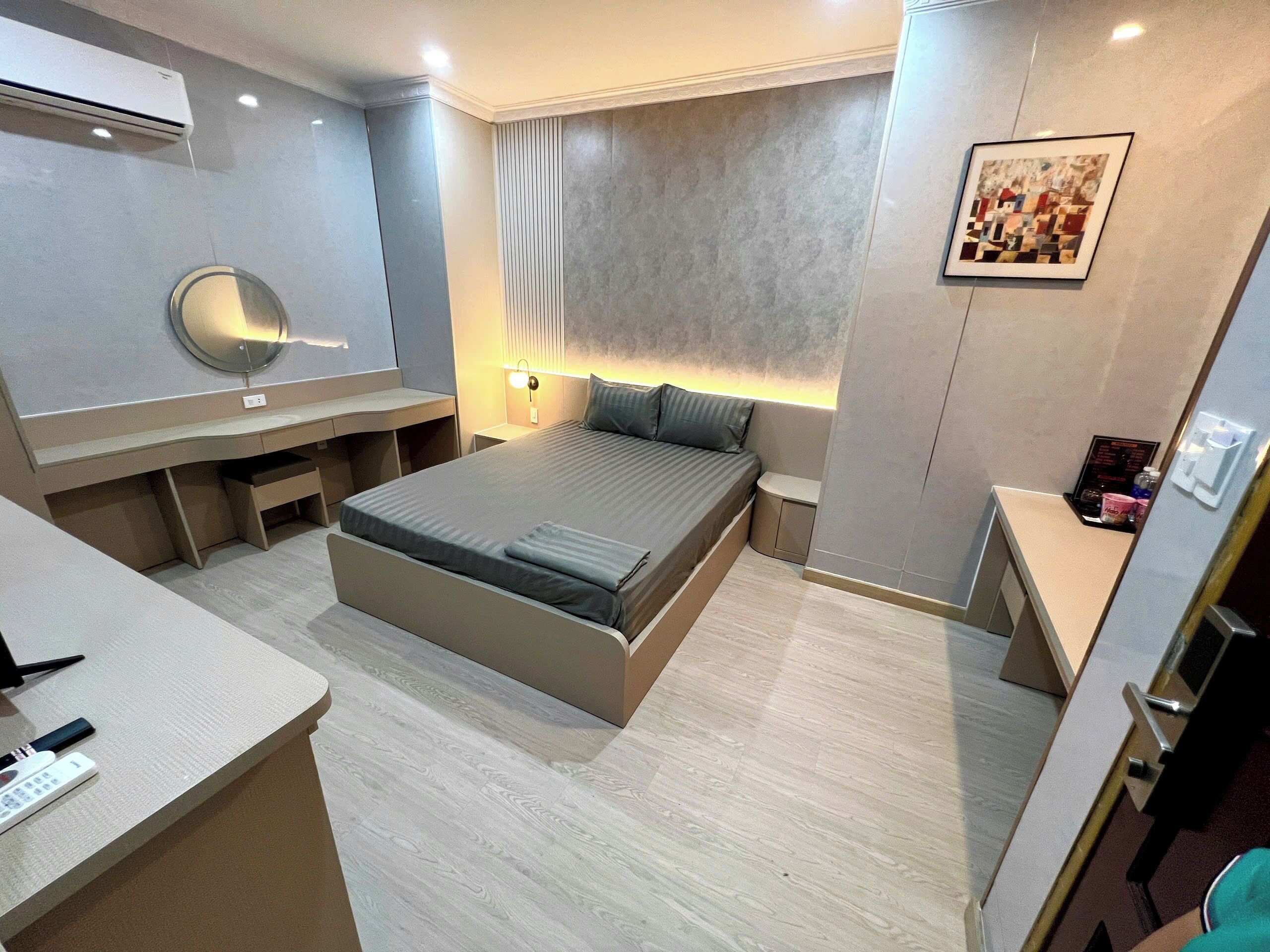 A modern hotel room with a bed, bedside tables, a desk, a chair, a mirror, and a wall-mounted artwork.
