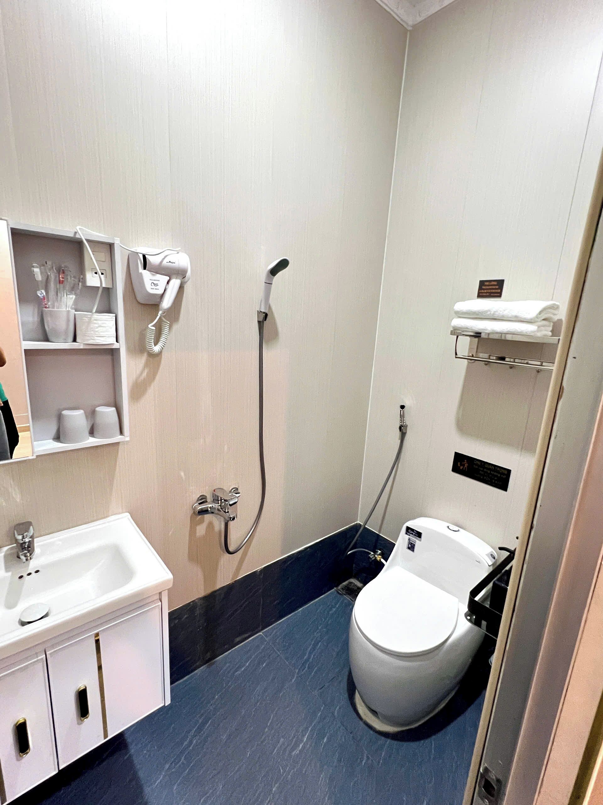 Ein kleines Badezimmer mit Waschbecken, Toilette und Dusche. Ein Föhn ist an der Wand angebracht und ein Ablagebrett bietet Handtücher.
