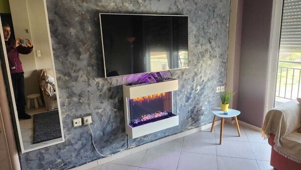Un salon moderne avec un mur à l’aspect pierre, un téléviseur à écran plat fixé au-dessus d’une cheminée, et une petite table ronde agrémentée d’une plante.