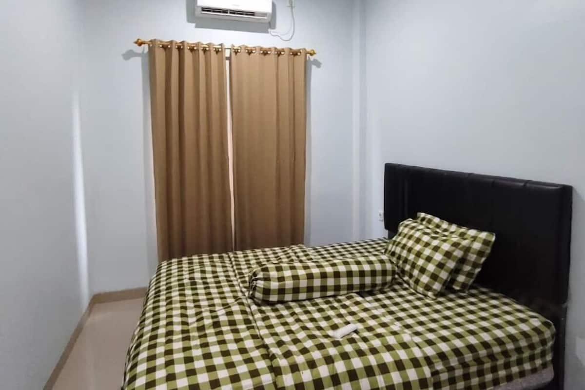 Una cama individual con una colcha a cuadros y almohadas a juego, un cabecero negro, cortinas color beige y un equipo de aire acondicionado en la pared.