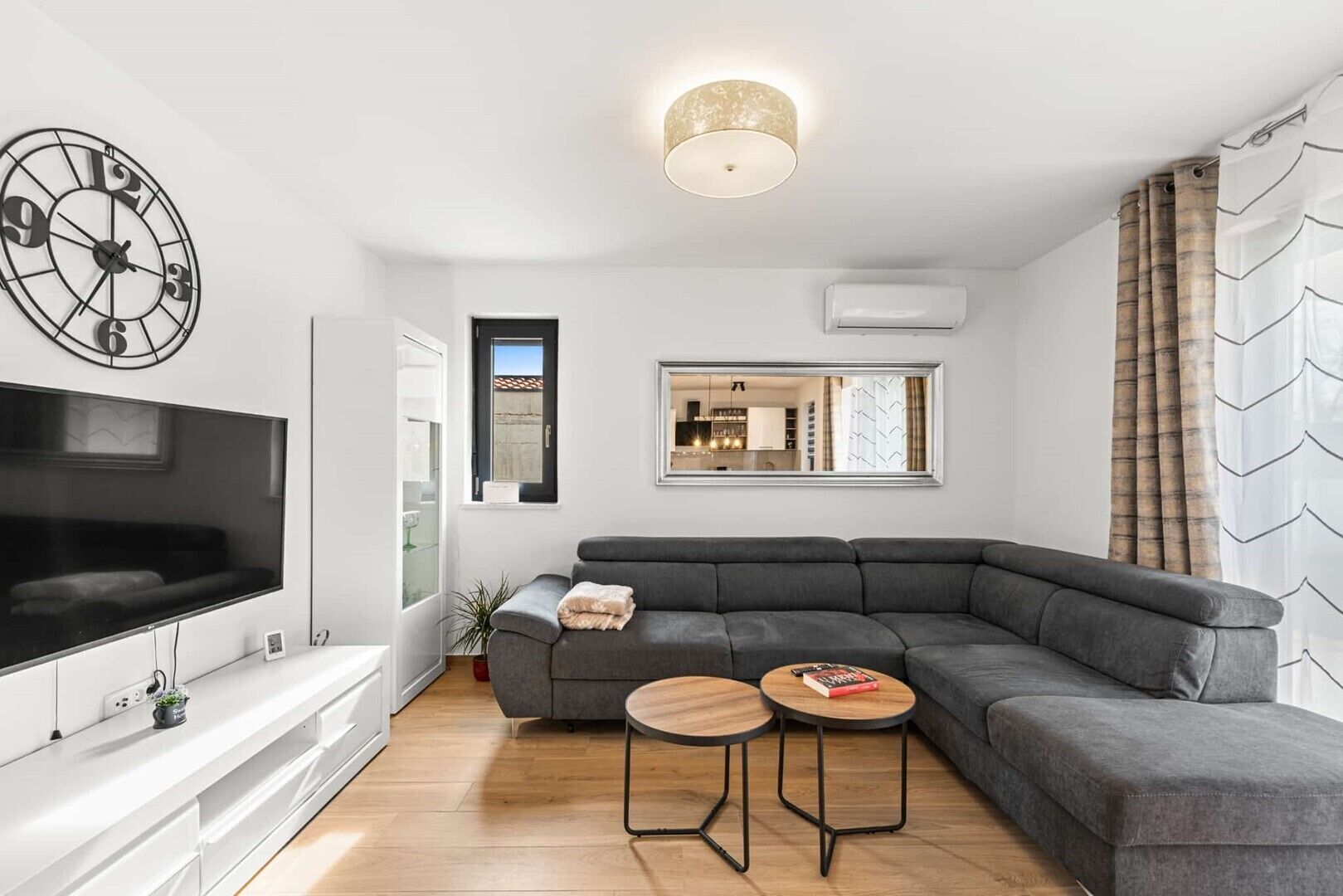 A modern living room with a sectional sofa, a flat-screen TV, a wall clock, and a mirror reflecting a kitchen area.