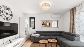 A modern living room with a sectional sofa, a flat-screen TV, a wall clock, and a mirror reflecting a kitchen area. - Apartment EM – Peaceful Stay near Poreč for 4 Guests (Poreč)