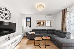 A modern living room with a sectional sofa, a flat-screen TV, a wall clock, and a mirror reflecting a kitchen area. - Apartment EM – Peaceful Stay near Poreč for 4 Guests (Poreč)