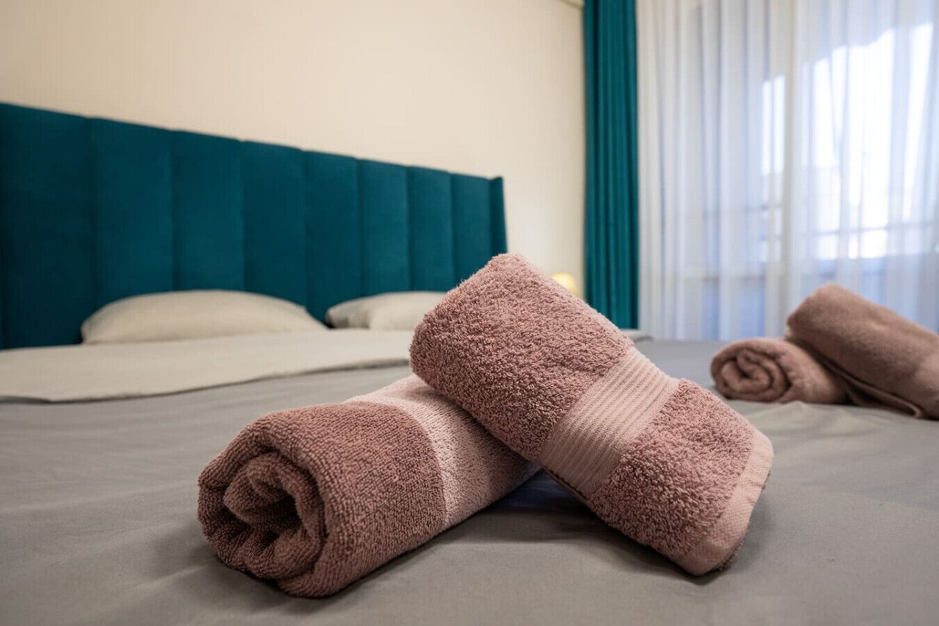A neatly made bed with a teal headboard, a folded pink towel, and a rolled-up towel on a grey bedspread.