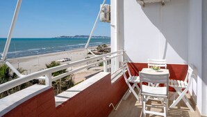 Un balcon avec des chaises blanches et une table, d’où l’on a une vue dégagée sur une plage, l’océan et le paysage urbain lointain.