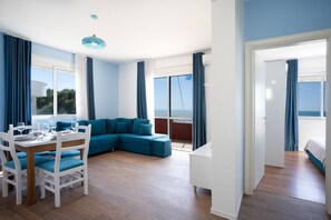 Ein modernes Wohnzimmer mit einem blauen Ecksofa, einem Esstisch mit Stühlen und großen Fenstern mit Meerblick.