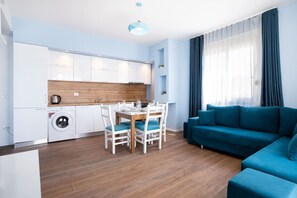 Eine moderne Wohnung mit Essbereich, einem Wohnzimmer mit blauem Sofa, einer Küche mit weißen Schränken und einer Waschmaschine.