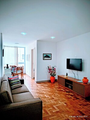Una sala de estar moderna con un sofá, un televisor, piso de madera y una zona de comedor con una mesa y sillas.
