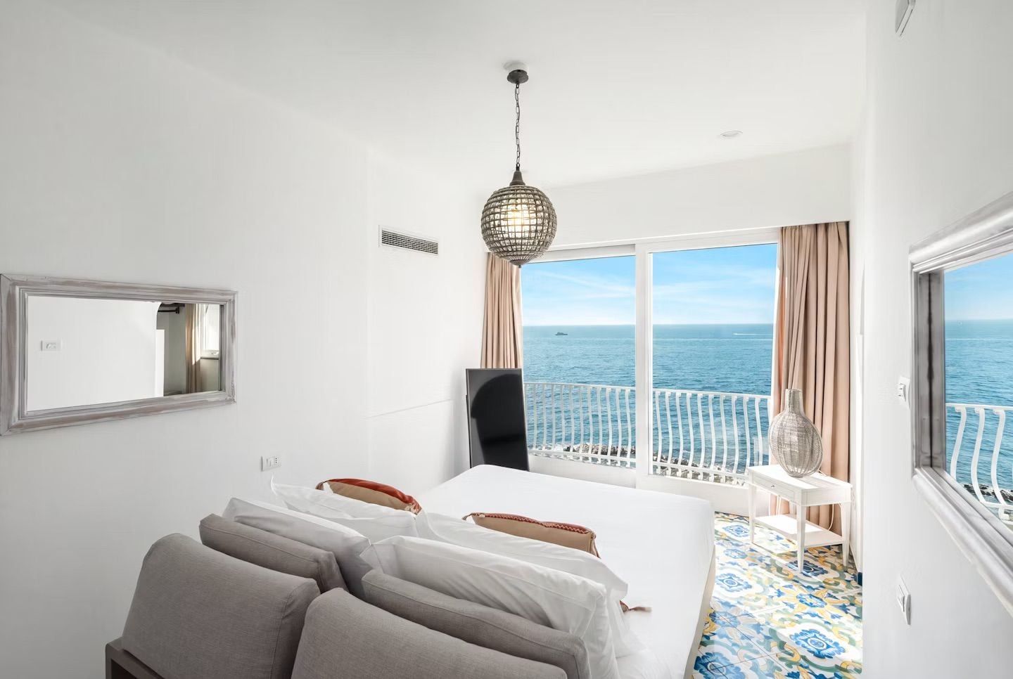 Una camera d'hotel con un letto, una scrivania, una sedia, un'ampia finestra vista mare e un lampadario.