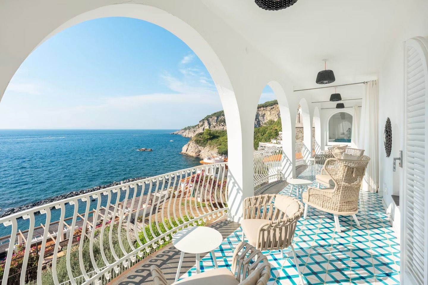 A balcony with a view of the sea, white furniture, and a patterned floor.