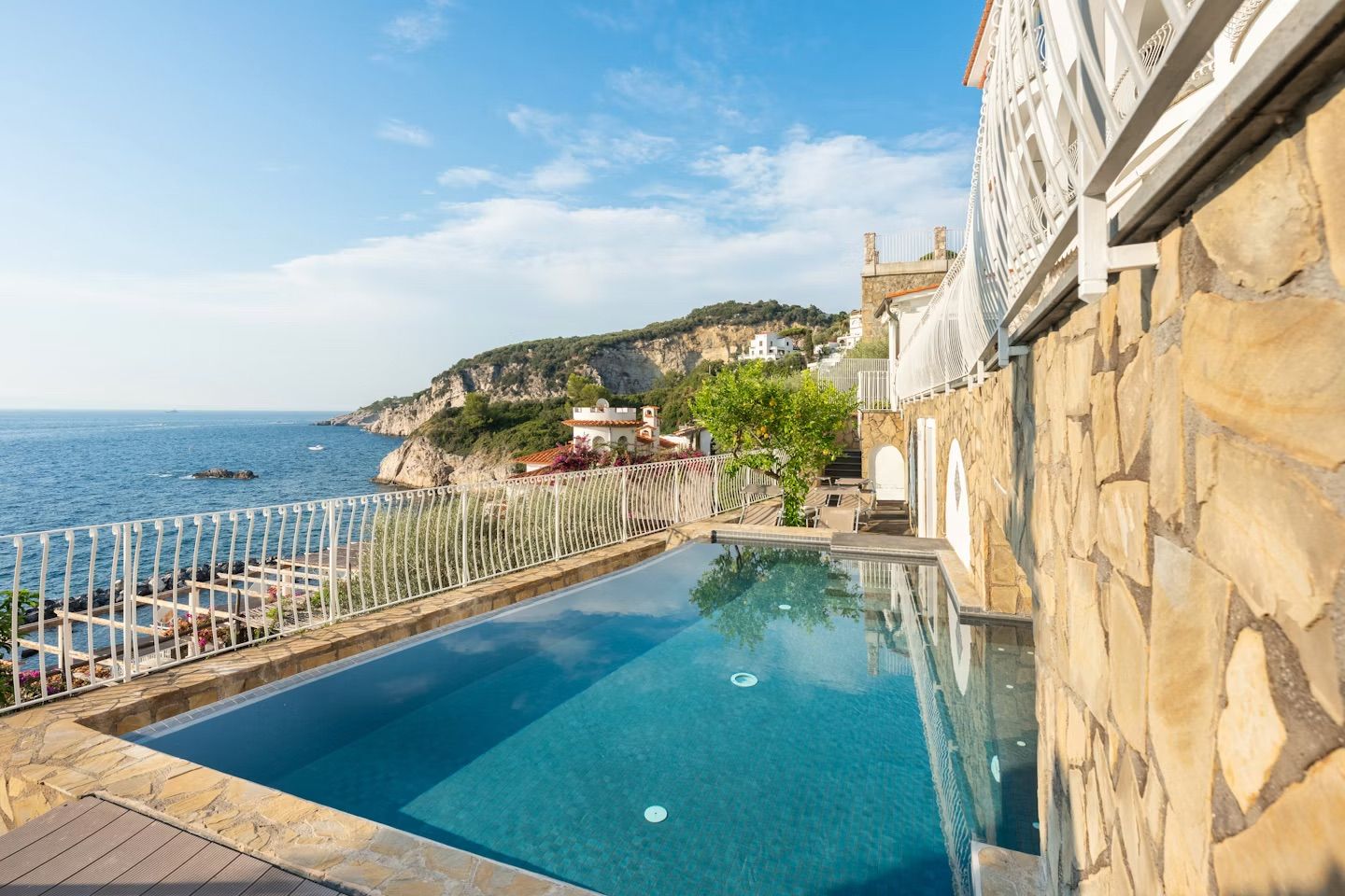 A clear blue pool with a stone wall on one side and a white railing on the other, overlooking a coastal landscape with buildings and cliffs.