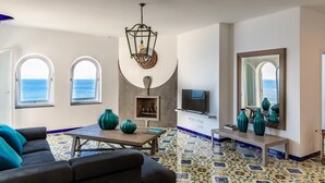 Una sala de estar con televisor, dos ventanas con vistas al océano, chimenea y suelo de baldosas.