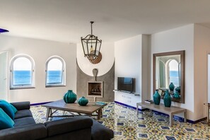 Ein Wohnzimmer mit Fernseher, zwei Meerblickfenstern, einem Kamin und einem Fliesenboden.