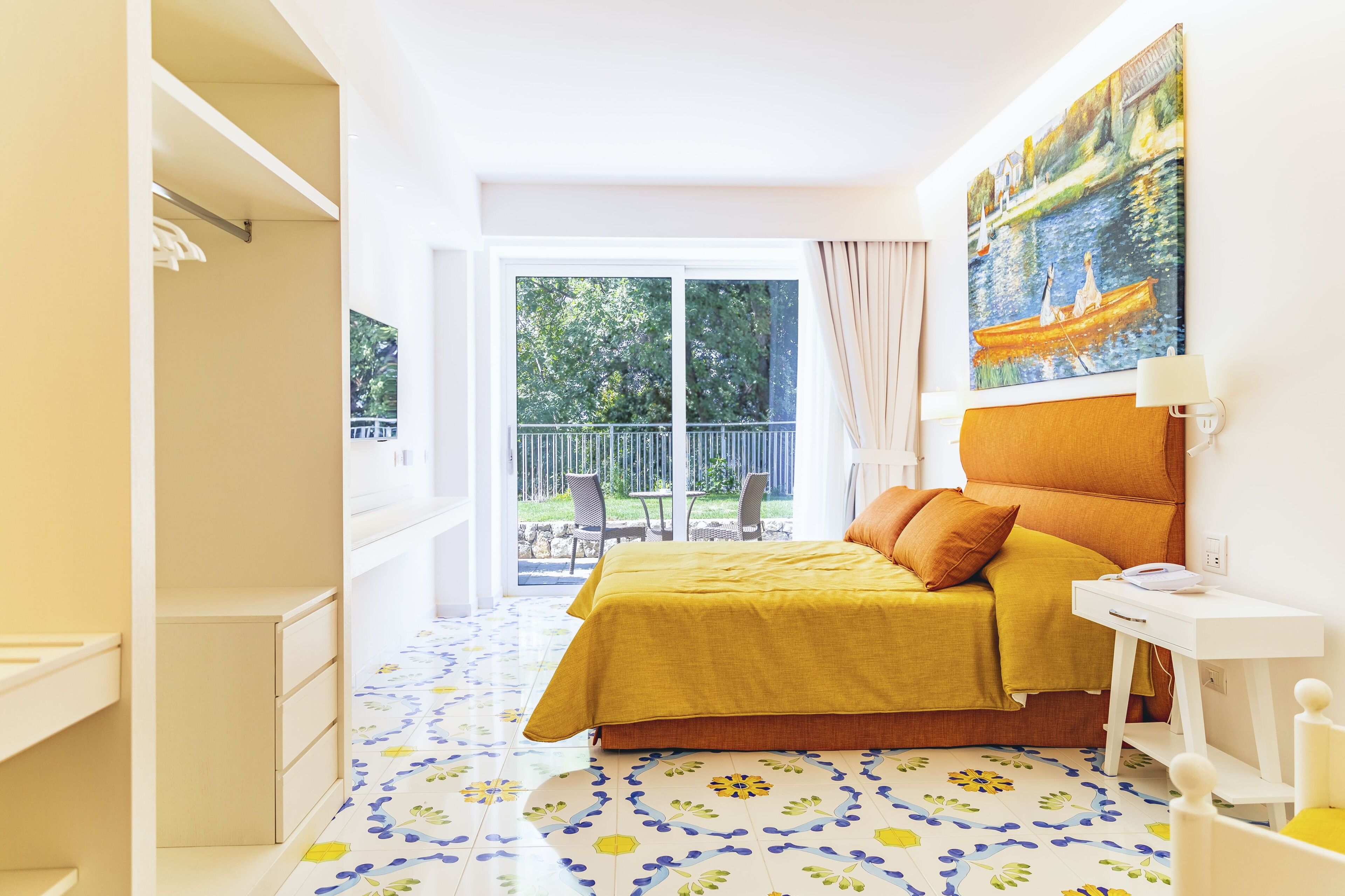 A bedroom with a bed, a desk, a chair, a painting, and a balcony with outdoor furniture.