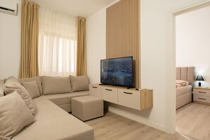 A modern living room with a beige sofa, a flat-screen TV mounted on a wooden cabinet, and a small bedside table with a lamp.
