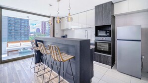 Una cocina moderna con una isla central, taburetes de bar, un refrigerador y vistas a edificios a través de ventanas amplias.