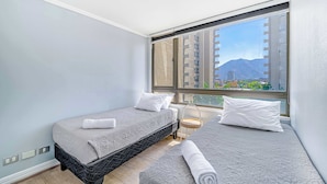 Una habitación de hotel con dos camas, una ventana con vistas a edificios y montañas, y una mesita con una lámpara.