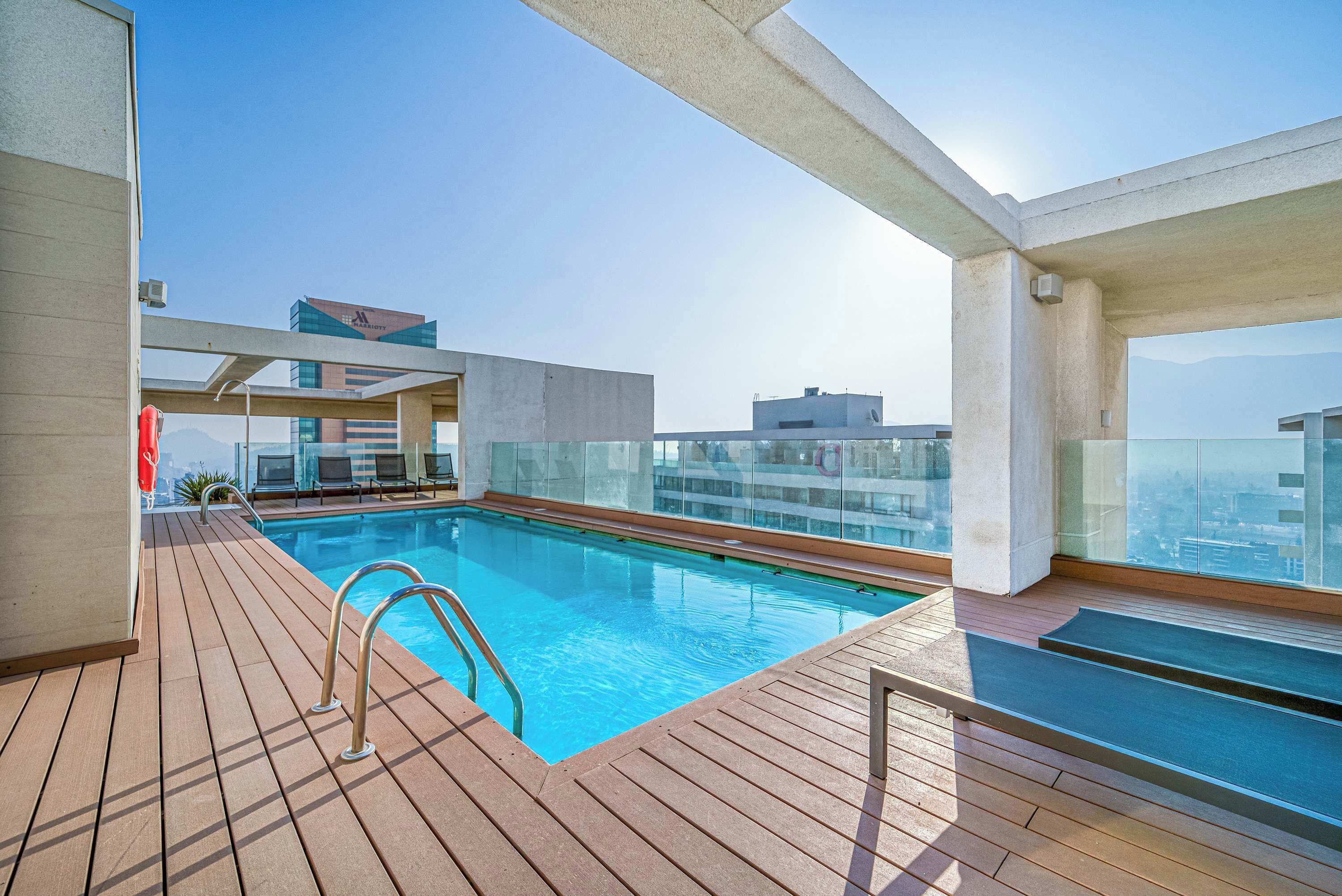 A rooftop swimming pool with a wooden deck, glass balustrade, and a clear view of the cityscape.