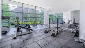 Un gimnasio con varios equipos de ejercicio, ventanas amplias y vistas a edificios y árboles.