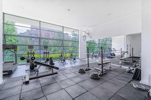 A gym with various exercise equipment, large windows, and a view of buildings and trees. - Brisa Capital 2BR - Wifi, Estacionamiento & Shopping Mall (Santiago)