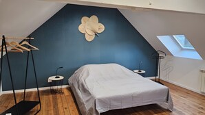 Un dormitorio con techo inclinado, una cama, un ventilador de pared, una mesita pequeña y un tragaluz.