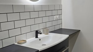 Un baño moderno con lavamanos blanco, tocador negro y un espejo redondo con luz.