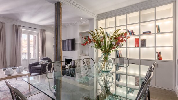 Un comedor moderno con una mesa de vidrio, sillas de metal, un florero con flores y una estantería con libros.