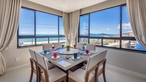 Un coin repas avec une table dressée pour quatre personnes, de grandes fenêtres avec des rideaux et une vue sur la plage et le paysage urbain.