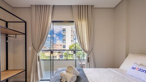 Une chambre d’hôtel avec un lit, une fenêtre avec des rideaux, une étagère et une vue sur des immeubles.