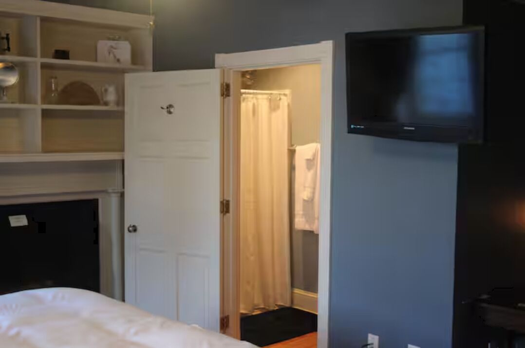 A hotel room with a bed, a television mounted on the wall, a door leading to a bathroom, and a shelf with various items. - Stock's Manor Bed and Breakfast #1 (Mechanicsburg)