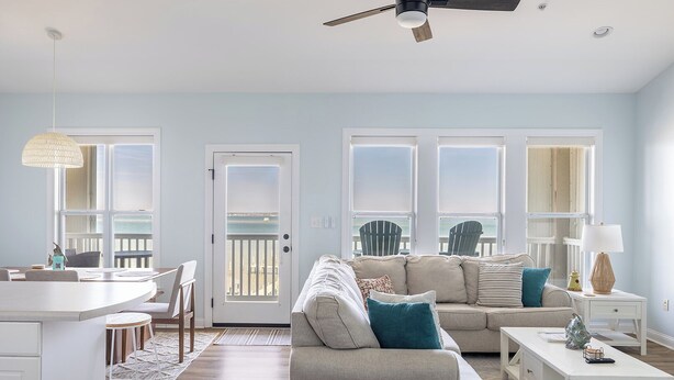 A modern living room with a sofa, coffee table, and dining area, featuring large windows and a ceiling fan. - Ocean Club N303, Soundfront Condo in Indian Beach! (Indian Beach)