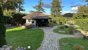 Un bâtiment rustique avec un toit de chaume, entouré d’un jardin bien entretenu, avec un sentier de pierres qui y mène.