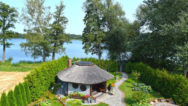Une maison à toit de chaume, dotée d’une terrasse circulaire en pierre, entourée d’une végétation luxuriante et offrant une vue sur un lac.