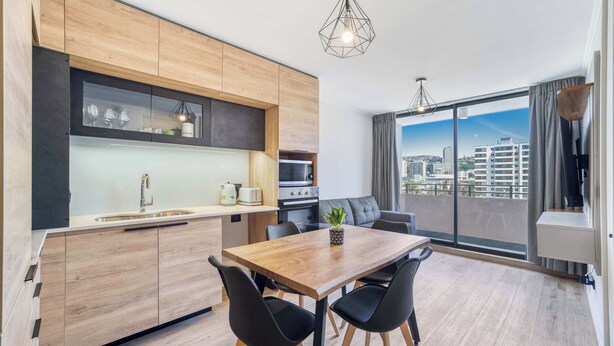 A modern kitchen with wooden cabinets, a dining table with black chairs, and a city view through large windows.