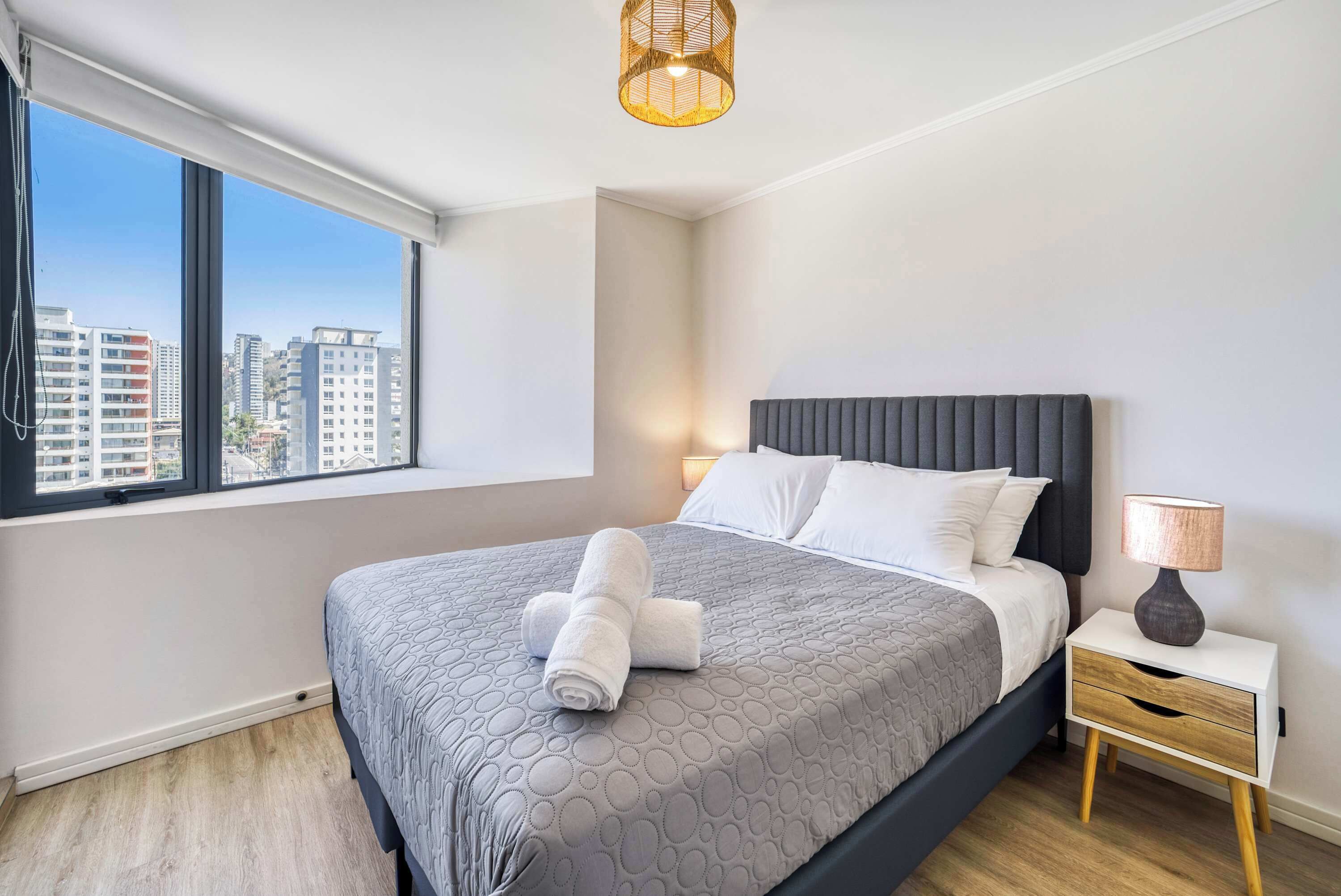 A modern hotel room with a large bed, a nightstand, a lamp, and a view of the city through large windows.