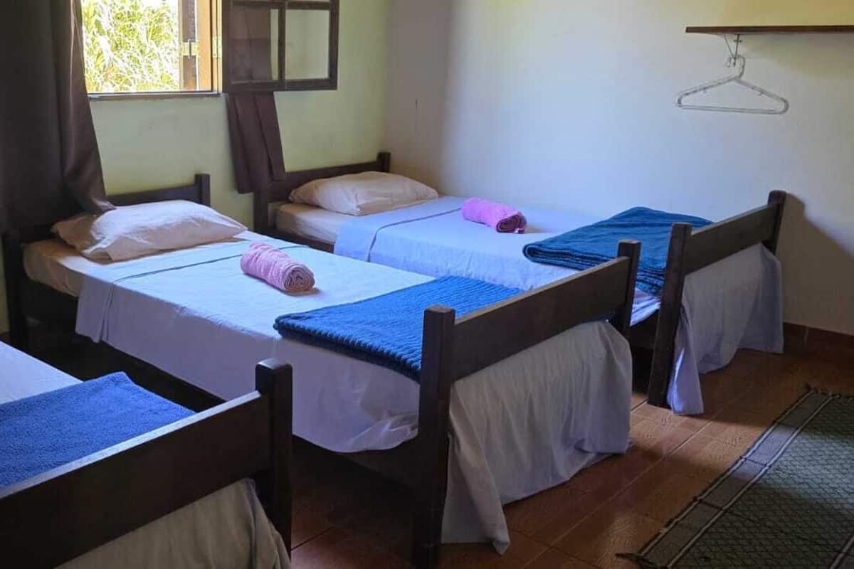 Une chambre avec deux lits simples, une fenêtre avec des rideaux, une tête de lit en bois et un porte-vêtements suspendu.