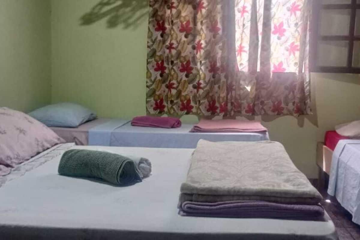 Une chambre avec deux lits, chacun étant équipé d’un oreiller et d’une couverture pliée. Il y a une fenêtre avec des rideaux à motifs floraux, ainsi qu’une petite table sur laquelle est posée une serviette rose.