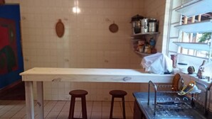 A kitchen with a long counter, two stools, a sink, and shelves with kitchenware. - O BREJO ENCANTADO HOSTEL E HOSPEDAGEM  (Embu das Artes)